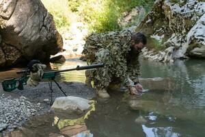 soldado en un camuflaje traje uniforme Bebiendo Fresco agua desde el río. militar francotirador rifle en el lado. foto