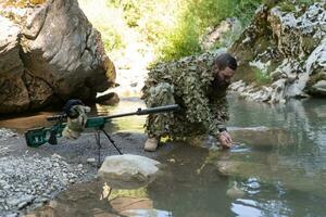 soldado en un camuflaje traje uniforme Bebiendo Fresco agua desde el río. militar francotirador rifle en el lado. foto