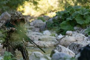 A military man or airsoft player in a camouflage suit sneaking the river and aims from a sniper rifle to the side or to target. photo