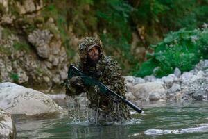 A military man or airsoft player in a camouflage suit sneaking the river and aims from a sniper rifle to the side or to target. photo