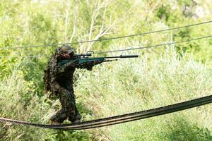 A military man or airsoft player in a camouflage suit sneaking the rope bridge and aims from a sniper rifle to the side or to target. photo