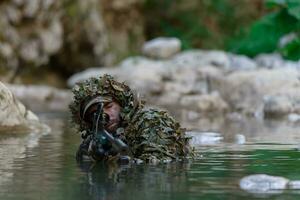 un militar hombre o airsoft jugador en un camuflaje traje furtivo el río y objetivos desde un francotirador rifle a el lado o a objetivo. foto