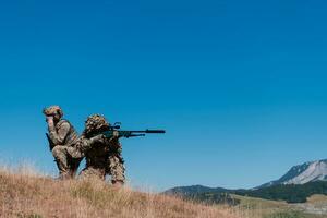 A sniper team squad of soldiers is going undercover. Sniper assistant and team leader walking and aiming in nature with yellow grass and blue sky. Tactical camouflage uniform. photo