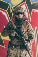 un barbado soldado en un especial efectivo uniforme soportes en frente de un dibujado pared con un rifle en su mano. selectivo atención foto