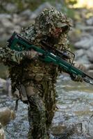 A military man or airsoft player in a camouflage suit sneaking the river and aims from a sniper rifle to the side or to target. photo