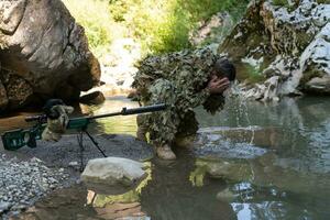 soldado en un camuflaje traje uniforme Bebiendo Fresco agua desde el río. militar francotirador rifle en el lado. foto