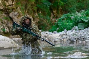 A military man or airsoft player in a camouflage suit sneaking the river and aims from a sniper rifle to the side or to target. photo