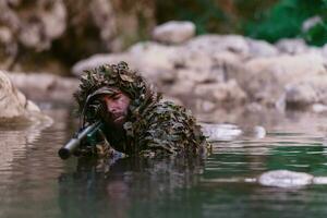 A military man or airsoft player in a camouflage suit sneaking the river and aims from a sniper rifle to the side or to target. photo