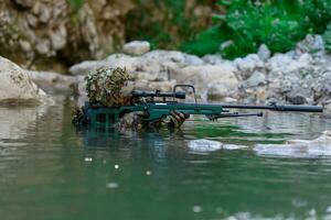 un militar hombre o airsoft jugador en un camuflaje traje furtivo el río y objetivos desde un francotirador rifle a el lado o a objetivo. foto