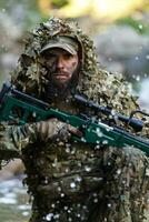 A military man or airsoft player in a camouflage suit sneaking the river and aims from a sniper rifle to the side or to target. photo