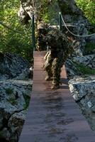 A military man or airsoft player in a camouflage suit sneaking the rope bridge and aims from a sniper rifle to the side or to target. photo