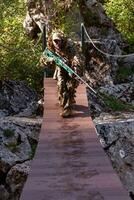 A military man or airsoft player in a camouflage suit sneaking the rope bridge and aims from a sniper rifle to the side or to target. photo