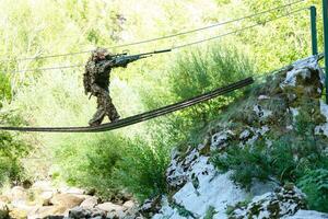 un militar hombre o airsoft jugador en un camuflaje traje furtivo el cuerda puente y objetivos desde un francotirador rifle a el lado o a objetivo. foto