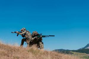 A sniper team squad of soldiers is going undercover. Sniper assistant and team leader walking and aiming in nature with yellow grass and blue sky. Tactical camouflage uniform. photo