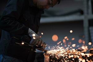 Heavy Industry Engineering Factory Interior with Industrial Worker Using Angle Grinder and Cutting a Metal Tube. Contractor in Safety Uniform and Hard Hat Manufacturing Metal Structures. photo