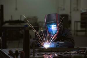 soldador profesional de la industria pesada que trabaja dentro de la fábrica, usa casco y comienza a soldar. enfoque selectivo foto