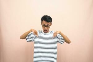 Asian man with glasses wearing casual striped shirt, whispering pose while pointing down. Isolated beige background. photo