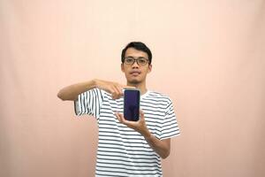 portrait of an asian man wearing glasses wearing a casual striped t-shirt. Shows smartphone screen with happy, surprised, happy expression, for promotion. Isolated beige background. photo