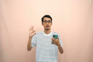 portrait of an asian man wearing glasses wearing a casual striped t-shirt. Posing showing thumbs up, recommending, okay, agreeing and holding smartphone. Isolated beige background. photo