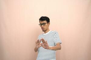 portrait of an asian man in glasses wearing a casual striped t-shirt. denote stop, cross prohibition sign, prohibit something, refuse,. beige background. photo