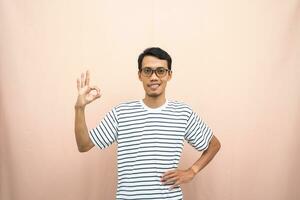 Asian man in glasses wearing casual striped shirt, posing giving thumbs up gesture or okay that agree. Isolated beige background. photo