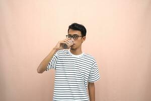 portrait of an asian man wearing glasses wearing a casual striped t-shirt. Enjoying drinking chocolate milk. Isolated beige background. photo