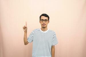 Asian man in glasses wearing casual striped t-shirt, pointing pose above empty space. Isolated beige background. photo