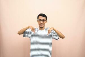 Asian man with glasses wearing casual striped shirt, whispering pose while pointing down. Isolated beige background. photo