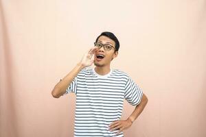 asiático hombre en lentes vistiendo casual a rayas camisa, gritos actitud con mano cerca boca, dando información. aislado beige antecedentes. foto