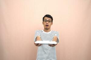 retrato de un asiático hombre en lentes vistiendo un casual a rayas camiseta. sorprendido a ver vacío platos. beige antecedentes. foto