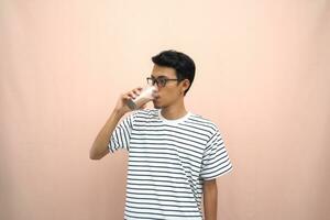 portrait of an asian man wearing glasses wearing a casual striped t-shirt. Enjoying drinking chocolate milk. Isolated beige background. photo