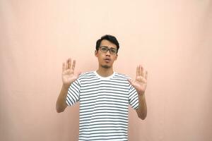 portrait of an asian man in glasses wearing a casual striped t-shirt. denote stop, cross prohibition sign, prohibit something, refuse,. beige background. photo