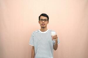 portrait of an asian man wearing glasses wearing a casual striped t-shirt. Enjoying drinking chocolate milk. Isolated beige background. photo