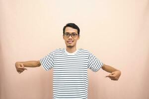 Asian man with glasses wearing casual striped shirt, whispering pose while pointing down. Isolated beige background. photo