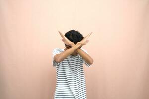 portrait of an asian man in glasses wearing a casual striped t-shirt. denote stop, cross prohibition sign, prohibit something, refuse,. beige background. photo