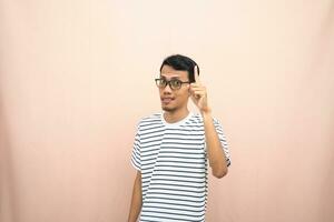 Asian man in glasses wearing casual striped t-shirt, pointing pose above empty space. Isolated beige background. photo