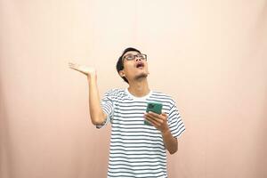 Asian man in glasses wearing casual striped shirt, pointing pose above empty space while holding smartphone. Isolated beige background. photo