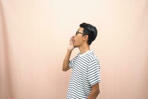 asiático hombre en lentes vistiendo casual a rayas camisa, gritos actitud con mano cerca boca, dando información. aislado beige antecedentes. foto