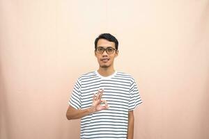 Asian man in glasses wearing casual striped shirt, posing giving thumbs up gesture or okay that agree. Isolated beige background. photo