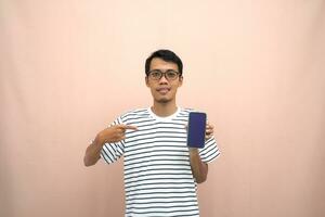 retrato de un asiático hombre vistiendo lentes vistiendo un casual a rayas camiseta. muestra teléfono inteligente pantalla con feliz, sorprendido, contento expresión, para promoción. aislado beige antecedentes. foto