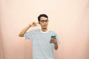 Asian man in glasses wearing casual striped t-shirt, thinking pose, looking for ideas while holding smartphone. Isolated beige background. photo