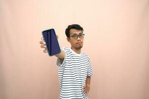 portrait of an asian man wearing glasses wearing a casual striped t-shirt. Shows smartphone screen with happy, surprised, happy expression, for promotion. Isolated beige background. photo