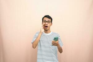 Asian man in glasses wearing casual striped t-shirt, whispering pose giving good news and surprise while holding smartphone. Isolated beige background. photo