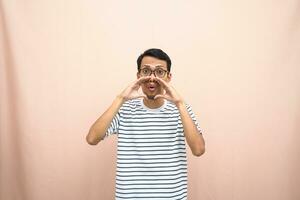 Asian man in glasses wearing casual striped shirt, shouting pose with hand near mouth, giving information. Isolated beige background. photo