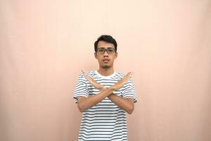 portrait of an asian man in glasses wearing a casual striped t-shirt. denote stop, cross prohibition sign, prohibit something, refuse,. beige background. photo
