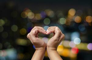 Female hand making heart symbol on night city colorful bokeh background. Love concept on Valentine day. photo