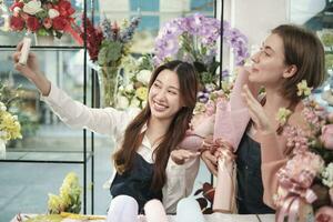 E-commerce business partner, two young female florist workers show floral arrangement, online live streaming, and selfies with smartphone application in bright flower shop, beautiful blossoms store. photo