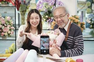 E-commerce business, Asian elder man and daughter florist shows floral arrangements and talks to camera via online live-streaming webcam application in a bright flower shop, beautiful blossoms store. photo