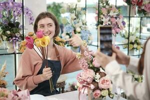 E-commerce business, one young White female florist demonstrates and shows floral arrangements via online live streaming with smartphone application in bright flower shop, beautiful blossoms store. photo