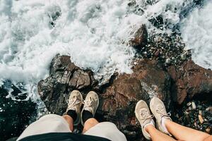 hembra y masculino piernas en pie por el agua en el rocas, en el costa. viaje foto. romántico calma atmósfera. agua espuma a rocoso rocas hermosa playa foto
